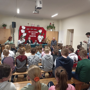 Prowadzący spotkanie poetyckiego z Marią Tarlagą i Zofią Solak w Szkole Podstawowej w Zaborowie. W tle dekoracja na czerwonym tyle biały napis. 